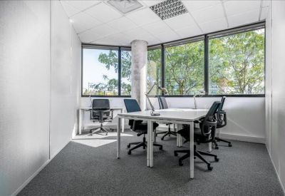 Bright corner office with multiple desks and large windows overlooking green trees.