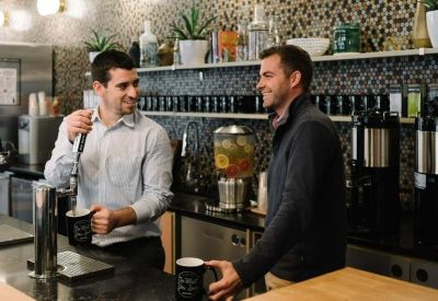 Close-up of a modern office kitchen area with two people talking and coffee machines.