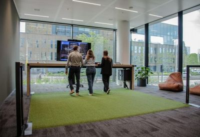 Three people working at a standing height long wooden table by large windows.