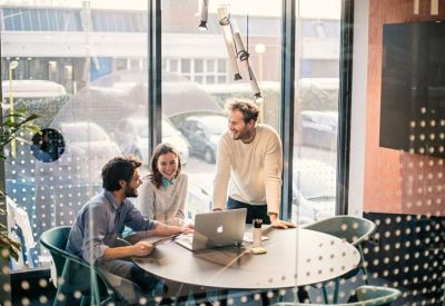 Colleagues collaborating at a round table with a laptop in a bright, glass-walled meeting space.