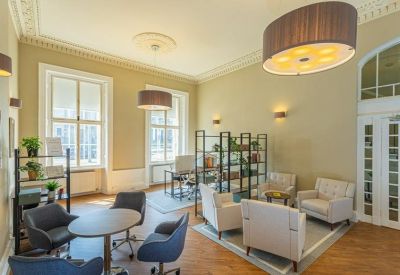 Bright communal lounge with grey and white armchairs, wooden floors, and large windows.