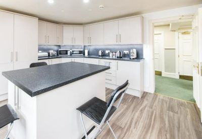 Modern communal kitchen area with a white island, breakfast stools, and wooden flooring.