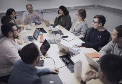 Collaborative meeting around a large wooden table with several laptops.