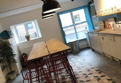 Modern communal kitchen and dining area with a long wooden table, red chairs, and geometric floor tiling.