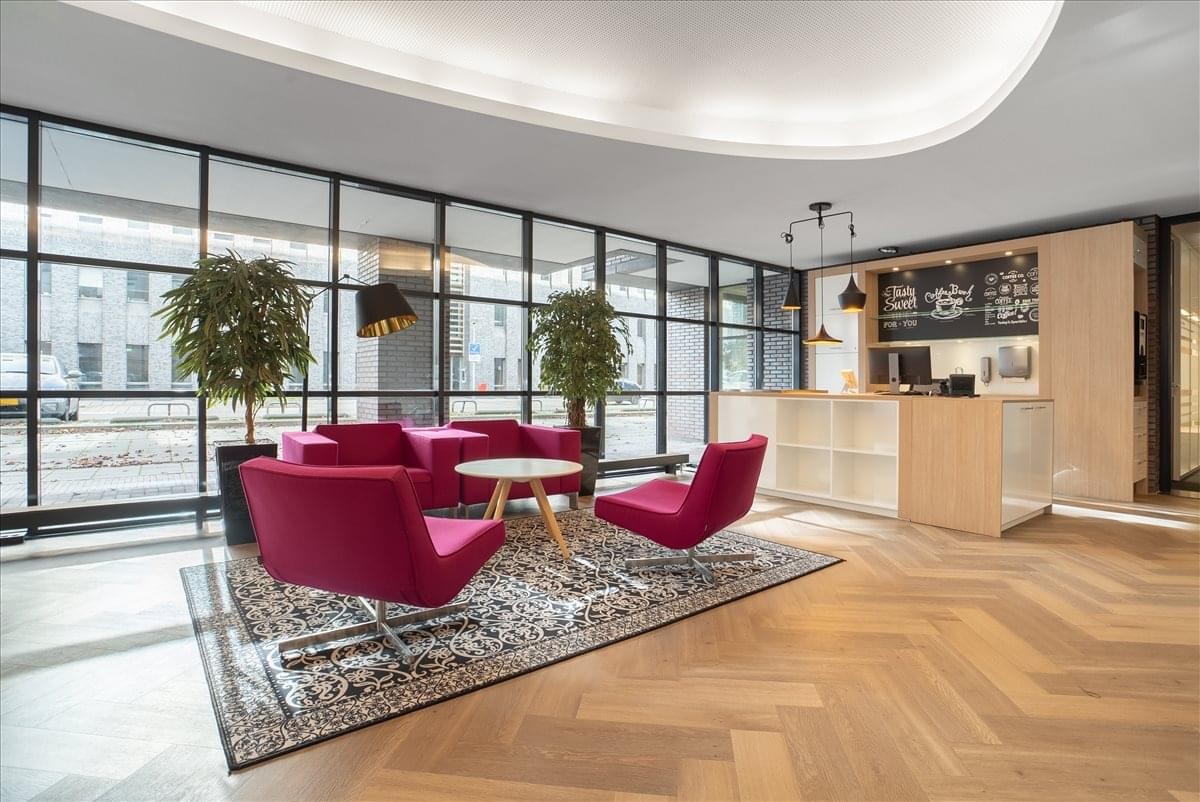Warm reception lounge with pink velvet armchairs on a patterned rug and herringbone wood flooring.