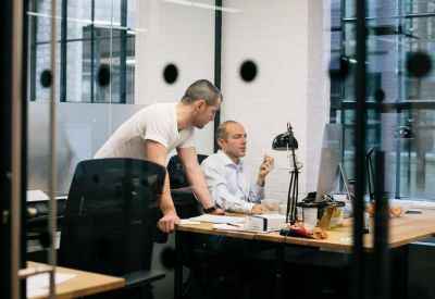Two people collaborating at a wooden desk in a modern glass-walled office suite.
