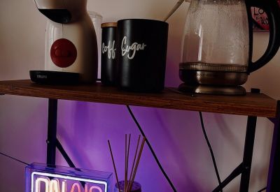 Coffee station with an espresso machine and neon sign on a wooden shelf.