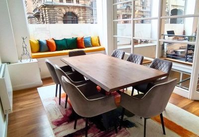 Bright dining area with a long wooden table, grey velvet chairs, and a colorful abstract rug.