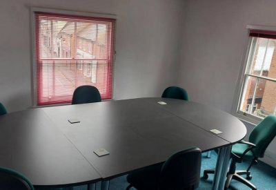 Large oval meeting table with teal chairs in a bright room with pink window blinds.