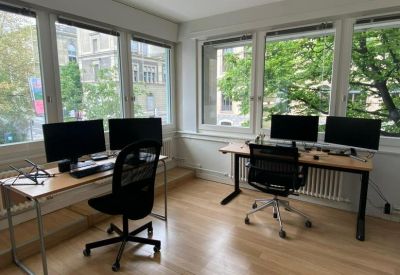 Bright workspace featuring two wooden desks, multiple monitors, and large windows overlooking green trees.