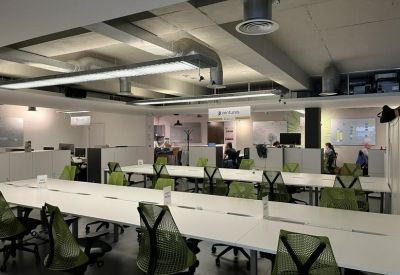 Modern open-plan workspace with rows of long white desks and ergonomic green chairs.