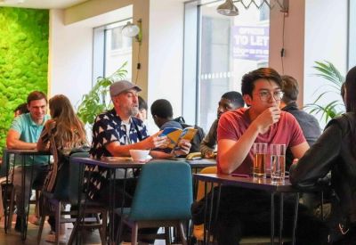 Lively coworking area with people working at small tables next to a green feature wall.