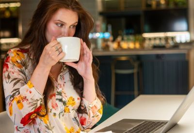 Woman working on a laptop while drinking coffee in a modern café setting.