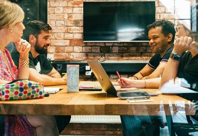 Meeting room with people collaborating around a large wooden table in front of a brick feature wall.