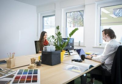 Open-plan office space with professionals working at a long wooden desk by large windows.