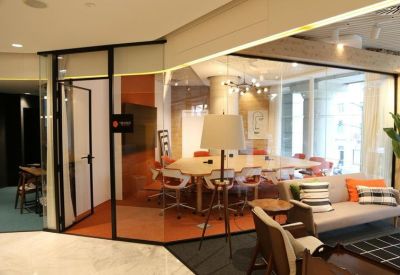 Glass-walled conference room viewed from a lounge area with a gray sofa and orange accents.