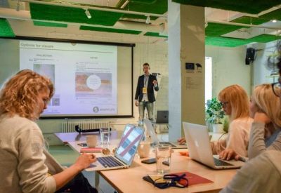 Meeting room setting with a presenter at a screen and attendees working on laptops at a wooden table.