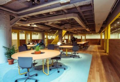 Sunlit open-plan office with round wooden desks and blue carpeting.