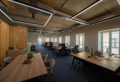 Spacious open-plan office with wooden desks, industrial ceilings, and large windows.