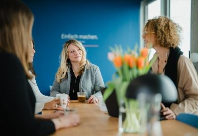 People chatting at a wooden communal table in a bright breakout area with a blue feature wall and fresh flowers.