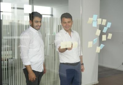Two professionals standing in a bright hallway using sticky notes on a glass partition.