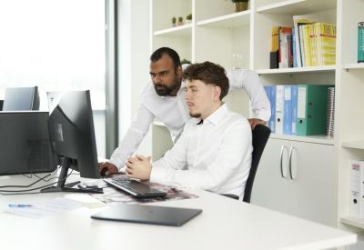 Two colleagues collaborating at a white desk with a dual monitor setup.