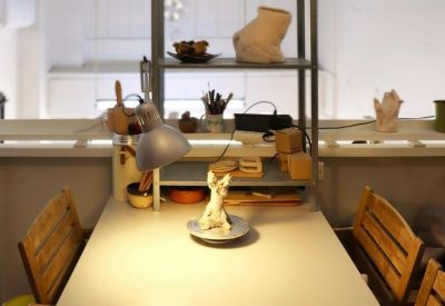 A long white communal work table under a tall industrial shelving unit holding ceramic sculptures.