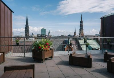 Rooftop terrace with wooden seating, potted plants, and views of city spires.