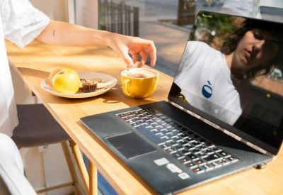 Close-up of a person at a laptop with a snack and coffee at a wooden cafe counter.