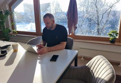A man working on a laptop at a white desk next to a bright window with a view of a snowy forest.