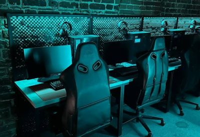 Gaming workstation row featuring high-back chairs and monitors against a brick wall with teal lighting.