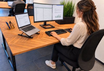 Spacious wooden desks in an open-plan office with a monitor, laptop, and indoor plants.