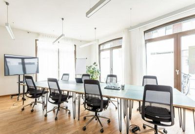 Spacious sun-drenched meeting room with a long table, black mesh chairs, and large windows.