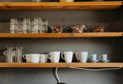 Communal kitchen area with wooden shelves holding mugs, glassware, and a coffee machine.
