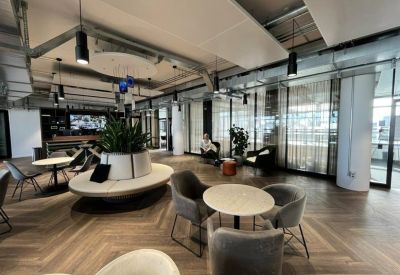 Spacious communal lounge with industrial ceilings, chevron wood floors, and modern circular seating around a large plant.