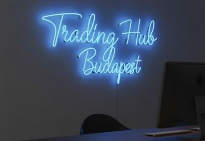 Blue neon sign on a grey wall reading Trading Hub Budapest above a dark wooden desk.