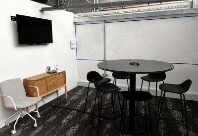 Meeting room with a high round table, black stools, and a wall-mounted TV screen.