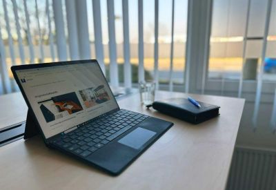 Laptop and notepad on a desk with natural light from window blinds.