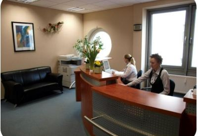 Bright reception desk with warm wood finishes, fresh flowers, and a black leather sofa.