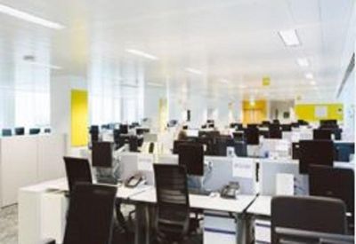 Open-plan office with white desks, ergonomic black chairs, and bright overhead lighting.