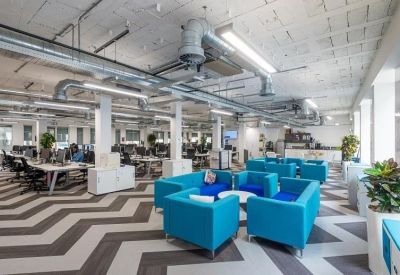 Spacious office interior with exposed ductwork, rows of desks, and bright blue armchairs.