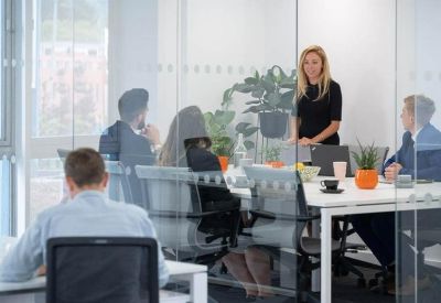 Glass-walled meeting room with people seated around a white table with orange accents.