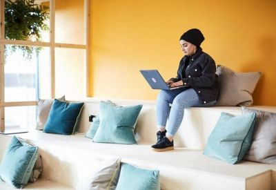 A person using a laptop on tiered seating with teal cushions in a bright lounge at NEO, Charlotte Street, Manchester.