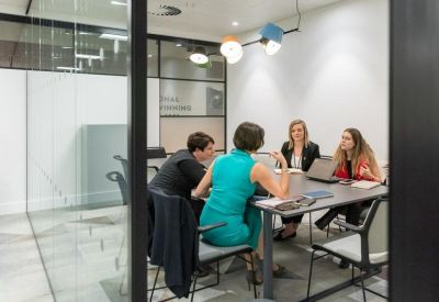 Meeting in progress around a dark conference table in a glass-walled room with colourful lighting.