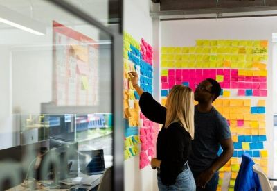 Two people standing by a vibrant glass wall covered in colourful sticky notes in a communal area.