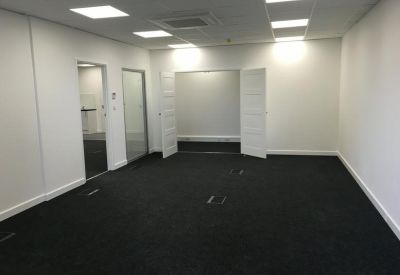 Large open-plan office space with black carpet and white walls leading to a secondary room.