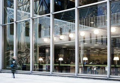 Glass-walled building interior showing a bright lobby area with warm lighting.