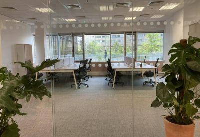 Bright open-plan office space with white desks and green potted plants viewed through a glass partition.