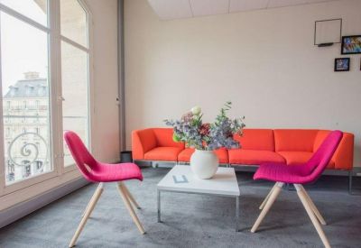 Bright breakroom featuring a bold orange sofa and pink designer chairs by a large window.