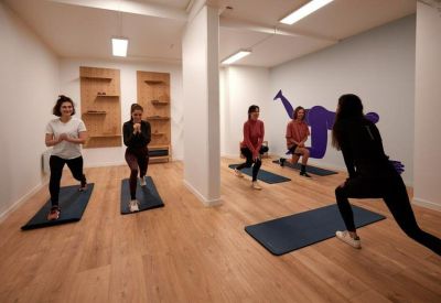 Bright fitness studio with wooden floors where people are performing lunges on yoga mats.
